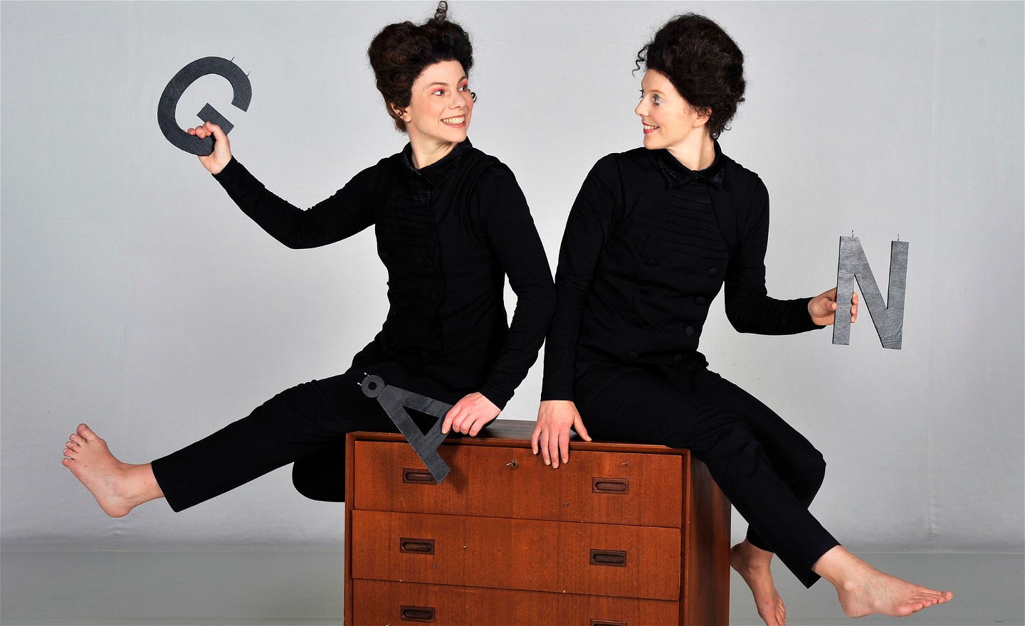 Sanna och Inger sitter på en byrå med ryggarna mot varandra och håller varsin bokstav, stora G och N.