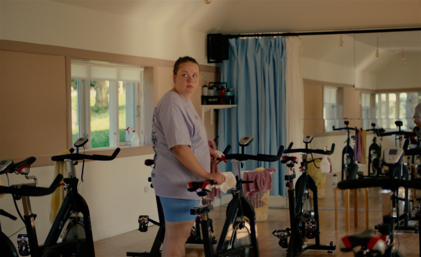 Tonårsflicka, klädd i short och t-shirt, håller i en träningscykel i ett rum fullt av cyklar och en spegelvägg.