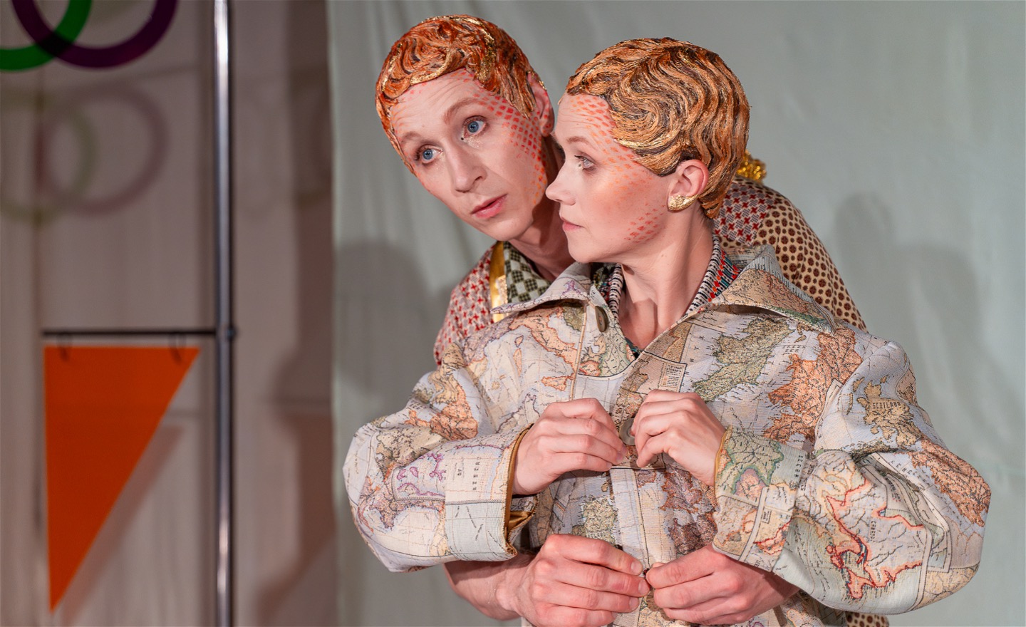 En dansare står och knäpper sin jacka. Hennes jacka har mönstret av en karta. Bakom henne står en annan dansare med sina armar runt henne och hjälper henne knäppa jackan. 