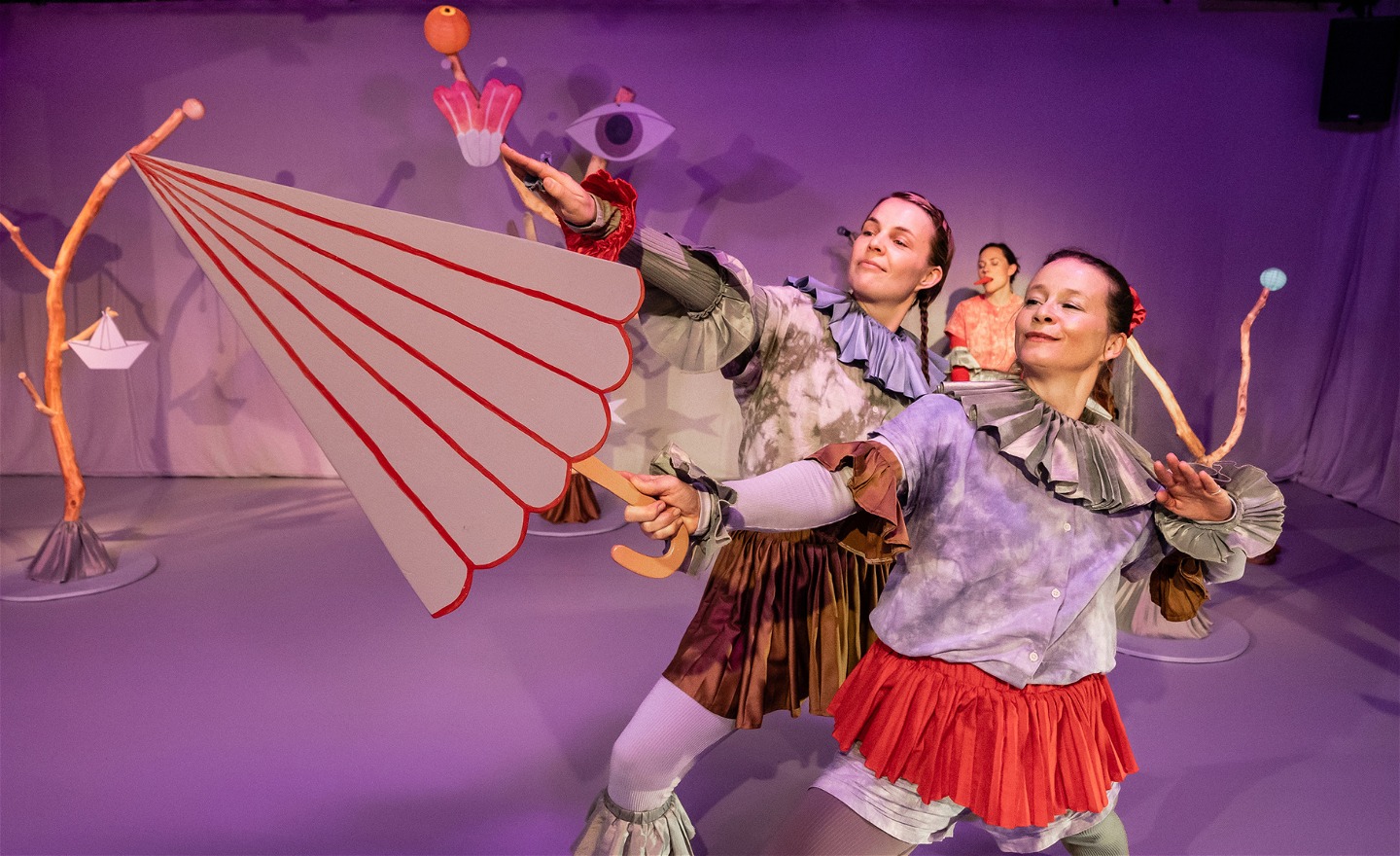 Dansarna Lisa och Åsa dansar med ett låtsas paraply. Färggranna figurer av trä i bakgrunden. 