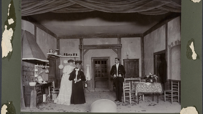 En interiörscen från ett herrgårdskök med två finklädda kvinnor och en betjänt.
