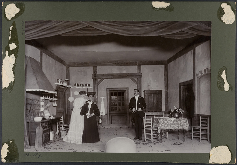 en interiörscen från ett herrgårdskök med två finklädda kvinnor och en betjänt.