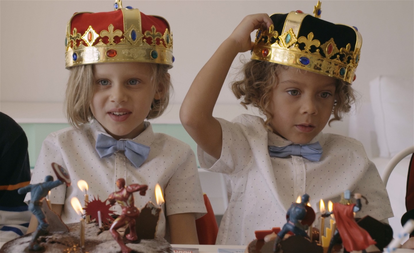 Tvillingarna sitter vid ett kalasdukat bord med tända ljus. Den ena har en guldkrona med rött tyg, den andre en guldkrona med blått tyg.