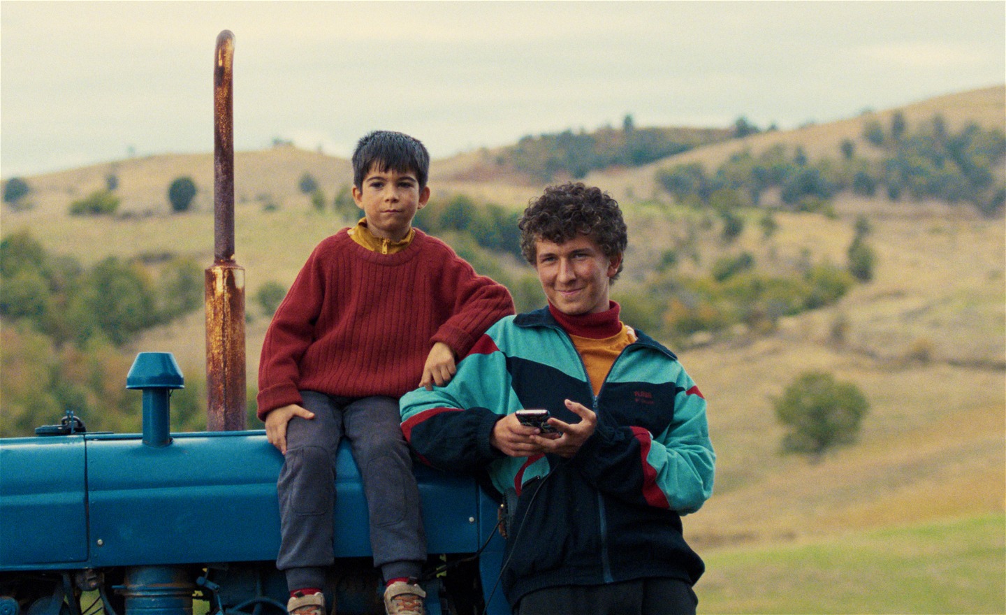 Tonårspojke står bredvid en blå traktor. Jämte honom sitter en yngre pojke som lagt sin arm mot hans axel. Bakom dem syns ett landskap med böljande kullar.