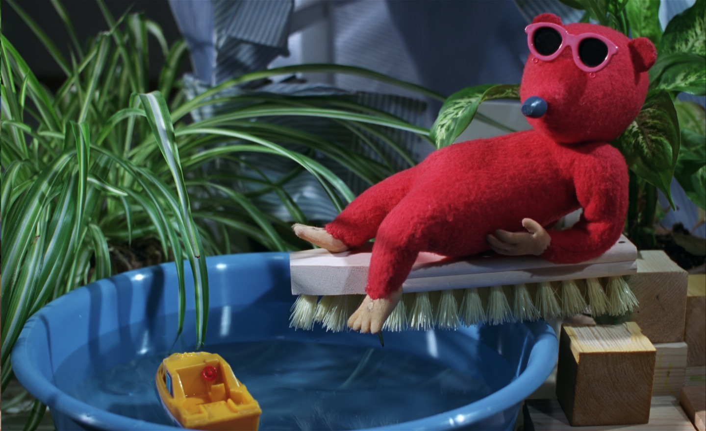 En röd figur med solglasögon sitter på en skurborste som balanserar på kanten av vattenfylld balja. I baljan ligger en gul plastbåt. I bakgrunden står två grönskande krukväxter.