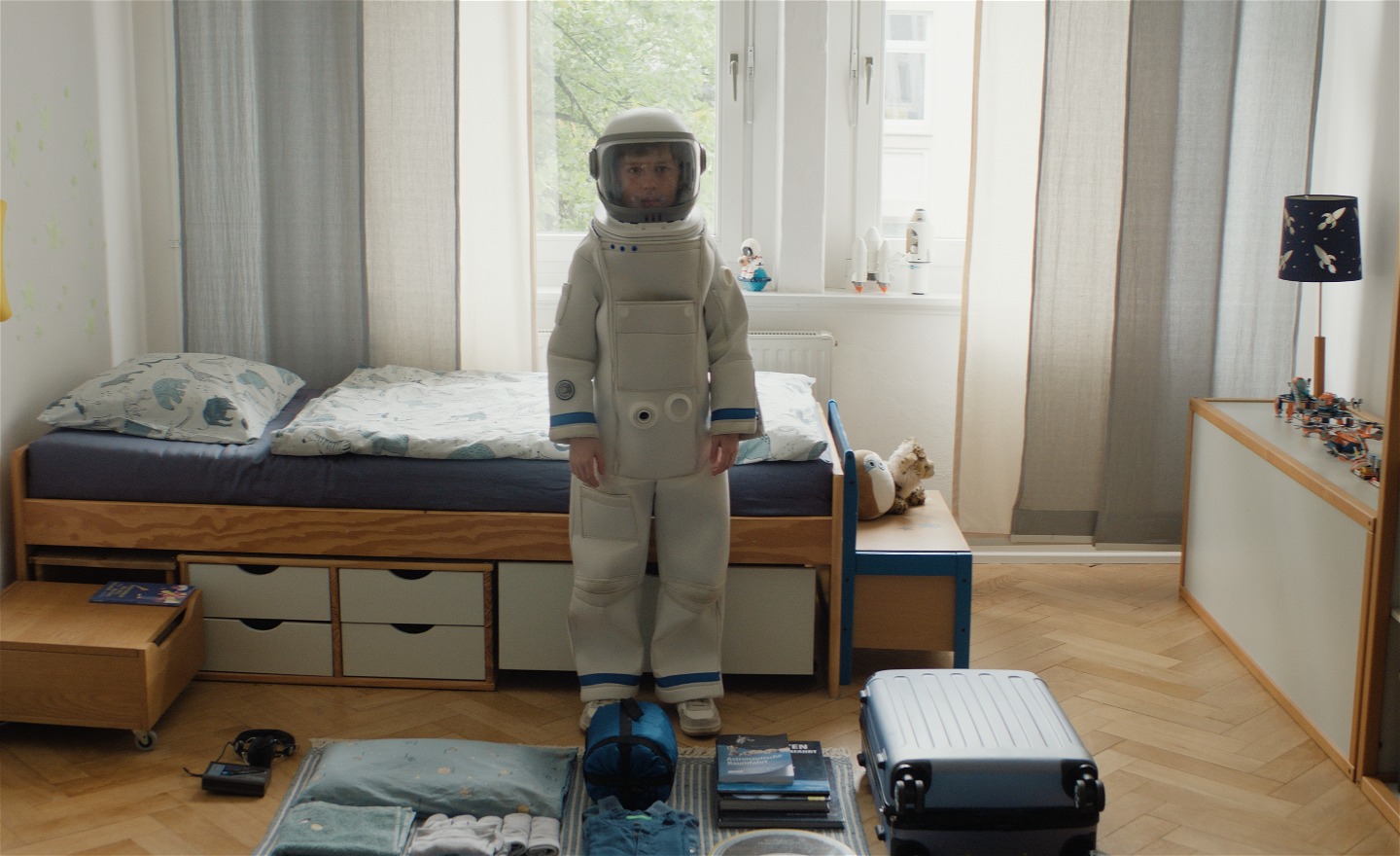 En pojke i astronautdräkt står framför en säng i ett pojkrum och ser in i kameran. Han ser allvarlig ut. 