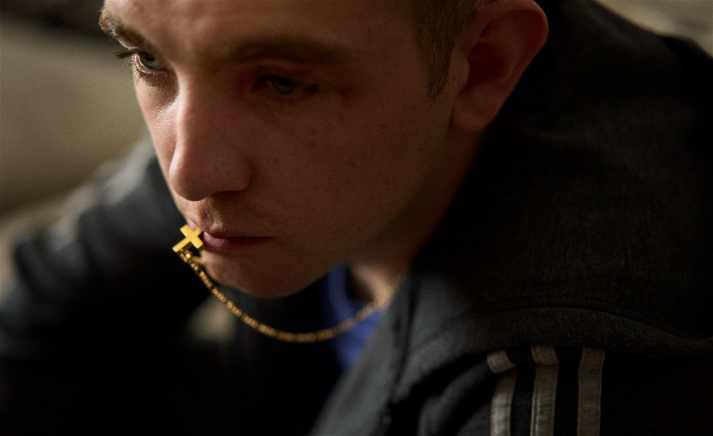 Ung man som ser allvarlig ut. I munnen håller han en del av ett hängsmycke från sitt halsband, ett guldfärgat kors.