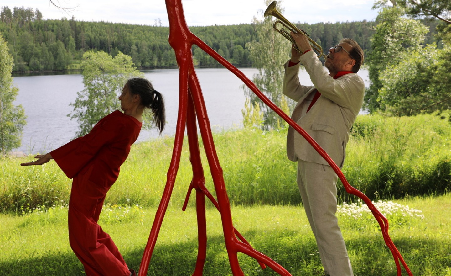 En rödklädd dansare och en trumpetare fångar formerna hos en röd trädskulptur med sina kroppar