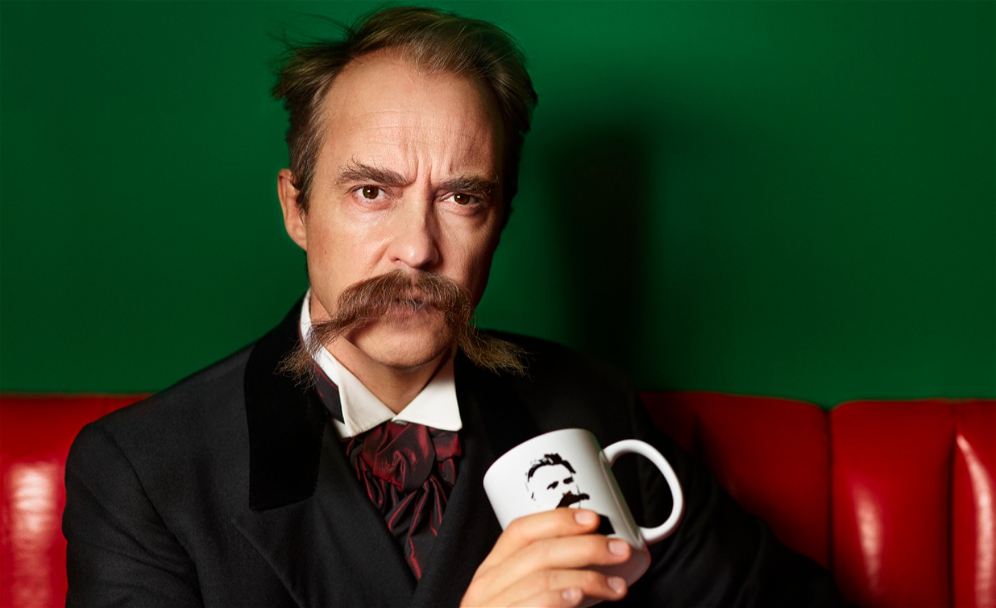 Jonas Karlsson som Friedrich Nietzsche med stor yvig mustasch och hållandes i en kaffekopp med ett porträtt av Nietzsche. 