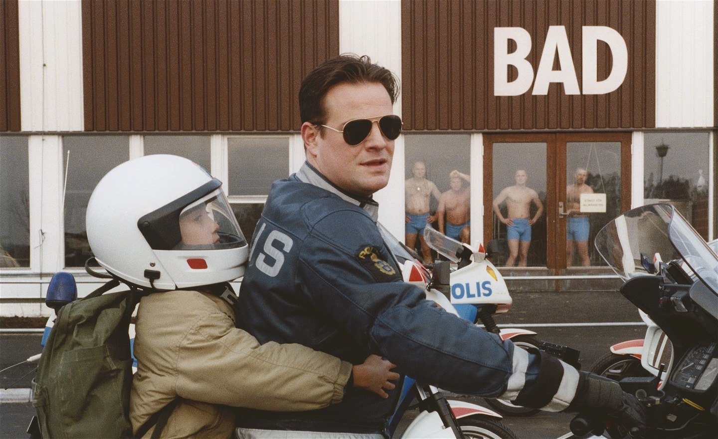 Man med solglasögon och polisuniform kör en polismotorcykel. Bakom honom sitter en pojke i beige jacka och vit motorcykelhjälm. Han håller armarna runt polisen som kör motorcykeln. I bakgrunden står fyra män iförda blå badbyxor och tittar på dem.