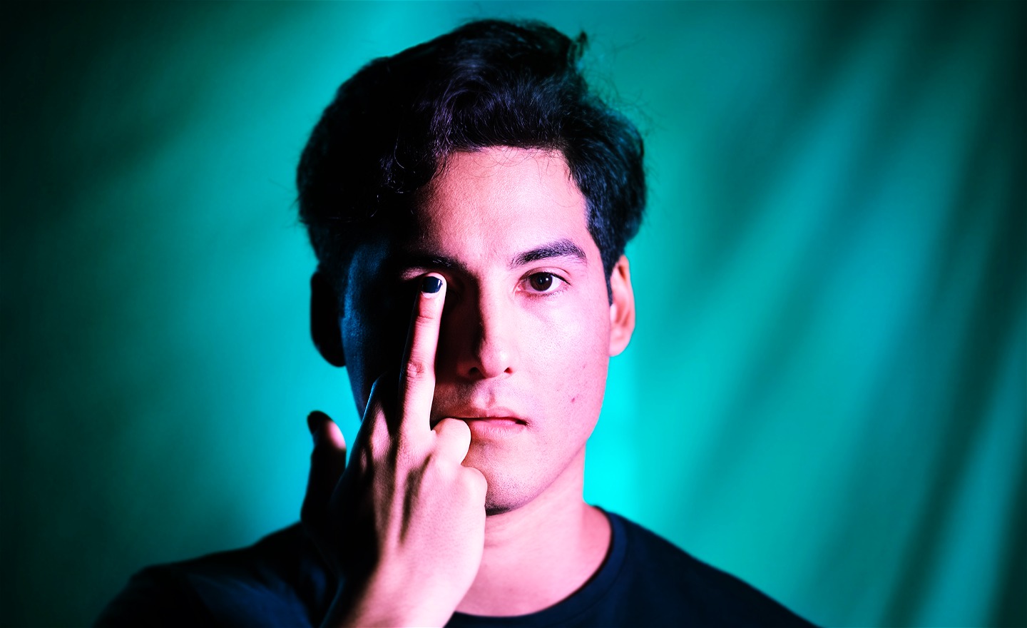 En person med mörkt hår står mot en blågrön bakgrund och håller upp sin hand med långfingret mot ena ögat; nageln är svartmålad.