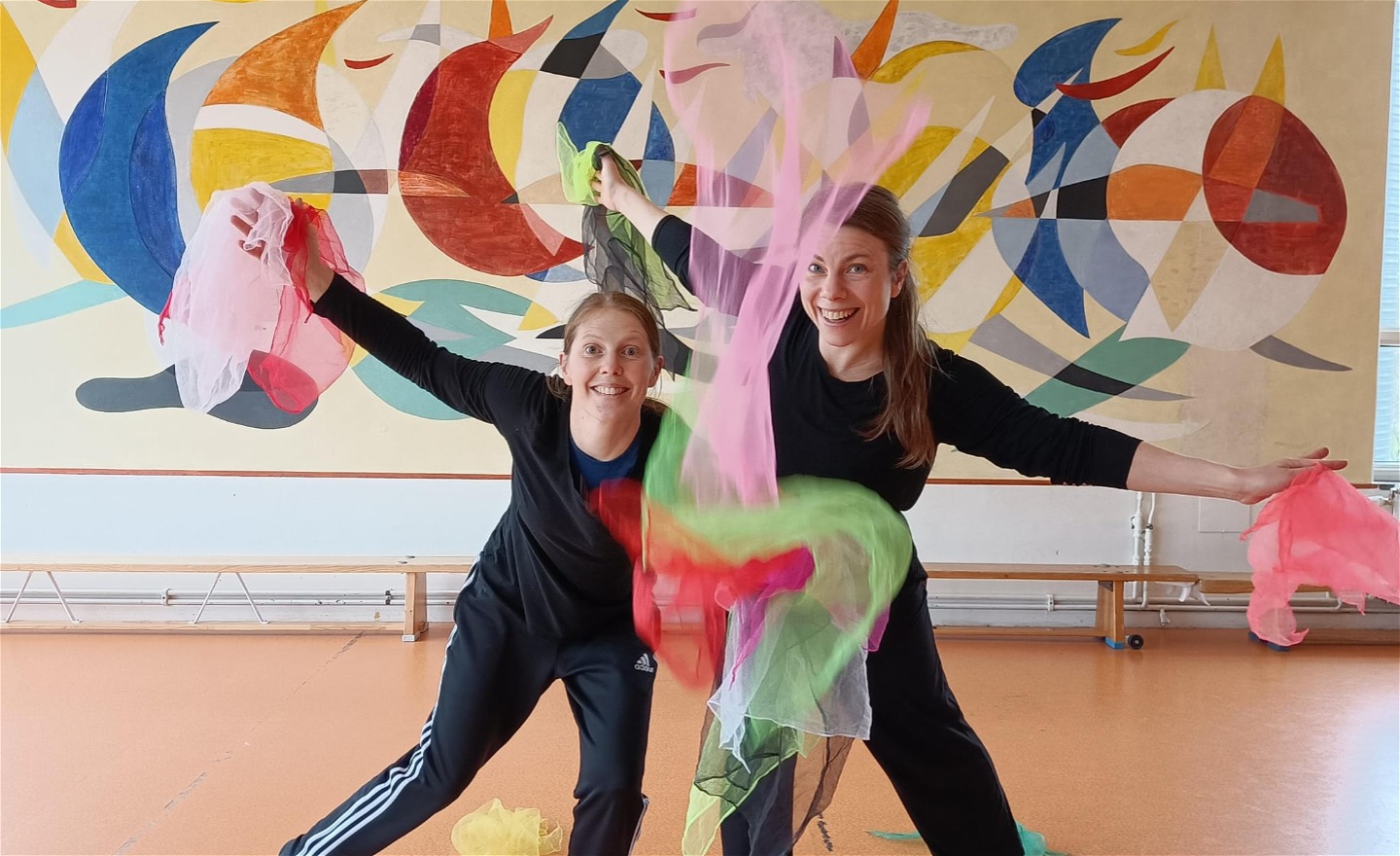 Två dansare ler mot kameran och svingar runt tunna sjalar i olika färger.