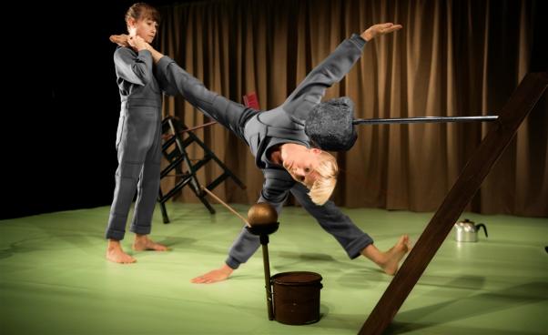 En akrobat balanserar en annan akrobat som bildar en skulptur med sin kropp. Det finns vardagsföremål i bilden som stege, kvast, burk och kaffekanna som är arrangerade som skulpturer.