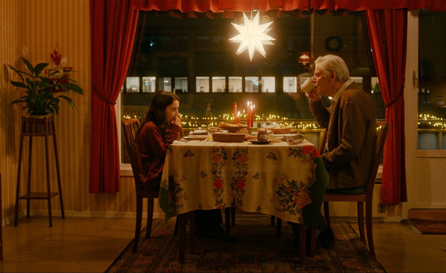 Flicka och gammal man sitter mittemot varandra vid frukostdukat bord med tänka juleljus. I fönstret hänger röda julgardiner och en adventsstjärna. Mannen dricker ur en mugg.