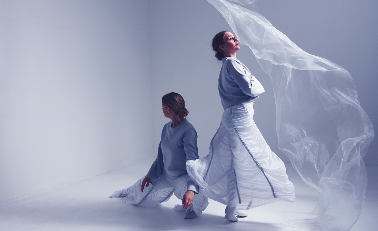 Två vitklädda dansare i ett vitt rum. Vitt skirt tyg faller till höger i bild