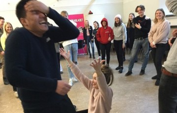 En person tar sig för pannan och skrattar, en person sitter nedanför och lyfter på armarna och en hel grupp står runt om och tittar på.