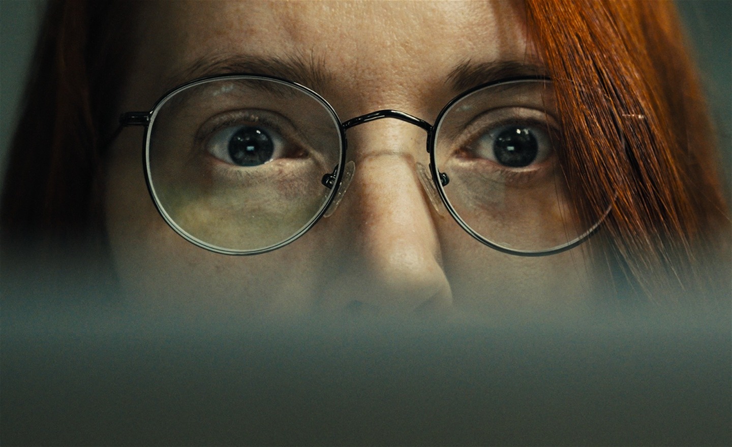 Kvinna med långt rött hår och svarta stålbågade glasögon tittar in i en datorskärm.