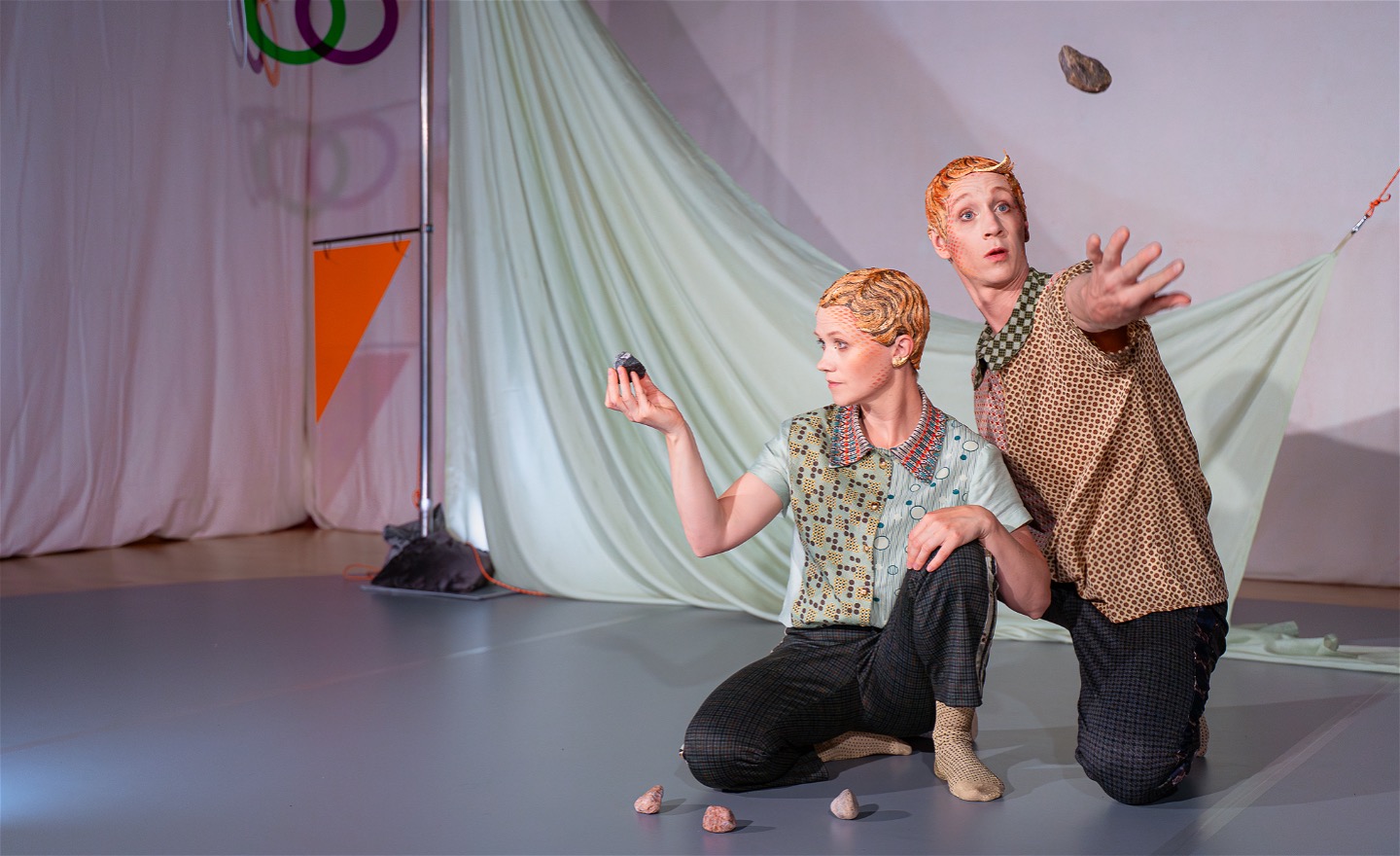 Två dansare i en scenmiljö där färgerna går i dämpat turkost, brunt, rosa och orange. Den ena dansaren håller upp en liten sten framför sig och tittar på den. Den andra bollar med en sten i luften.