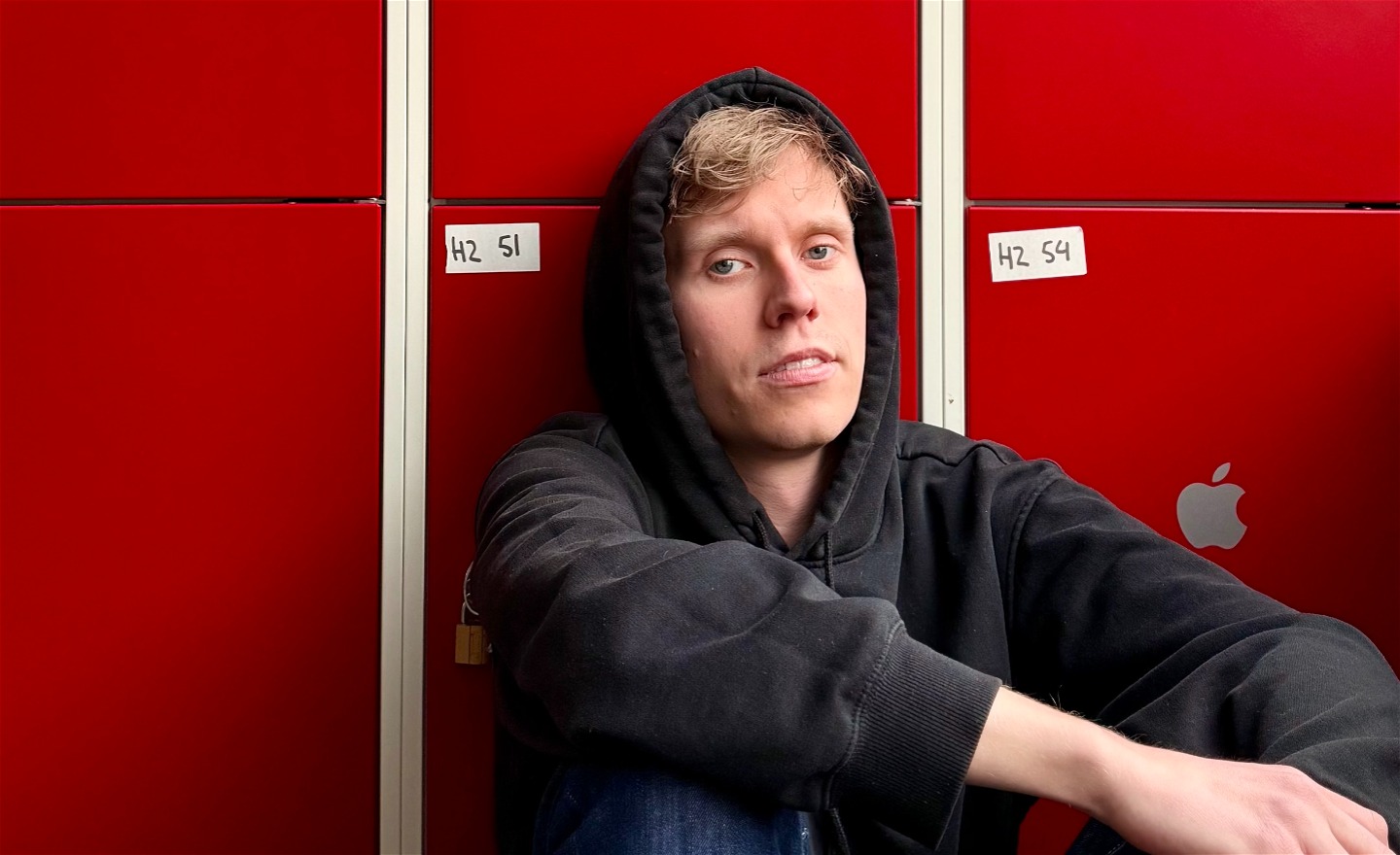 En pojke i huvtröja sitter framför röda skolskåp och tittar mot oss