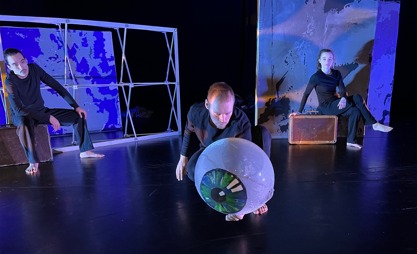 Tre dansare i svart, två sitter på resväskor, en håller ett stort öga i handen