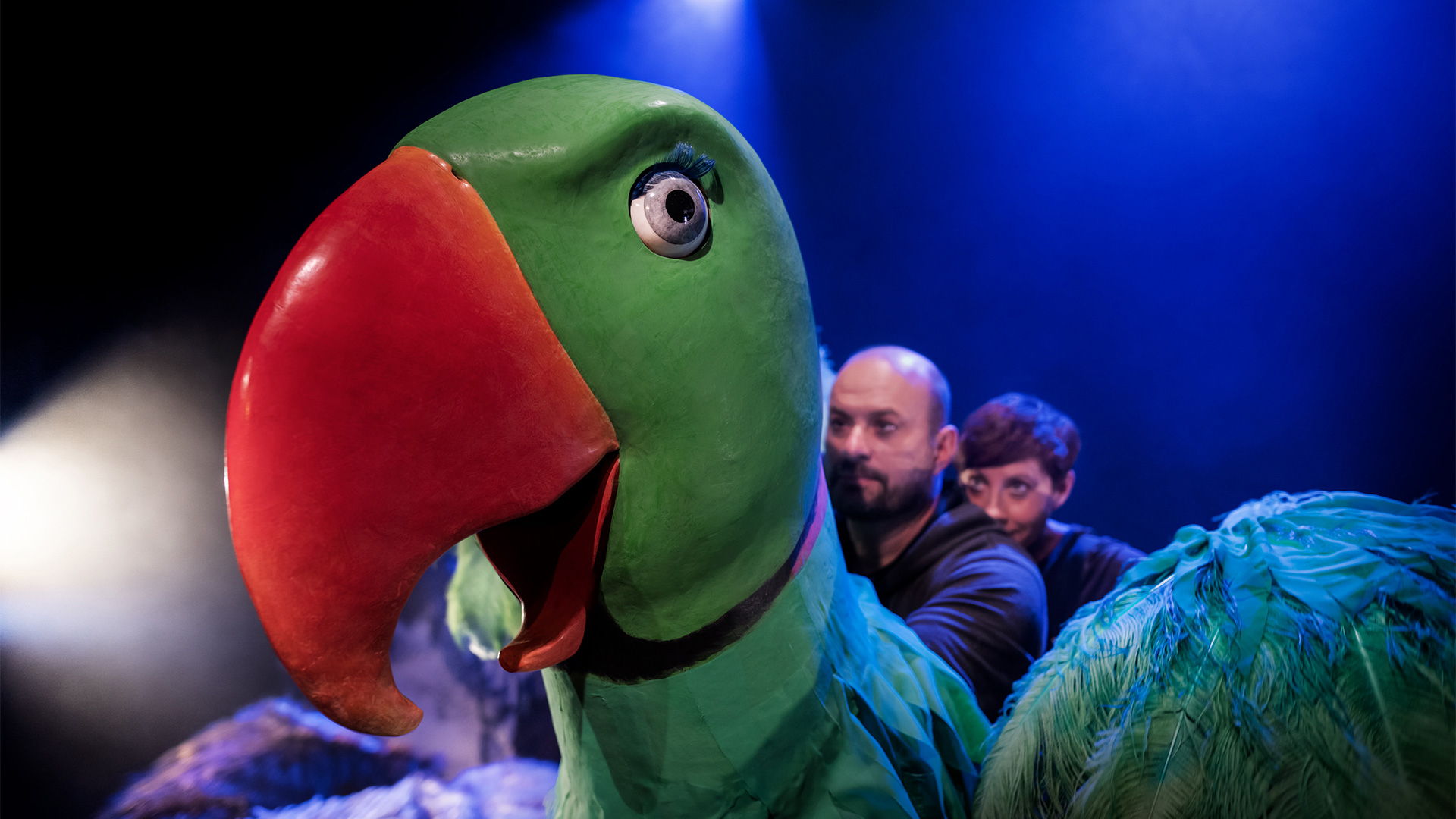 en marionettdocka i form av en stor grön fågel med orange näbb samt två personer bakom som styr dockan.