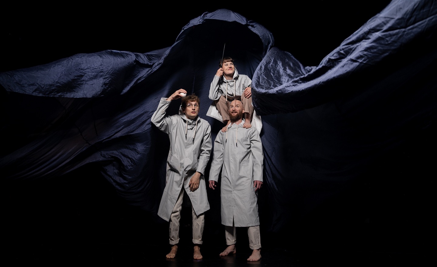 Tre artister i grå regnrockar står framför ett blått, böljande tyg. En sitter på en annans axlar och håller i ett paraply. En annan står bredvid och håller upp en jongleringsboll.