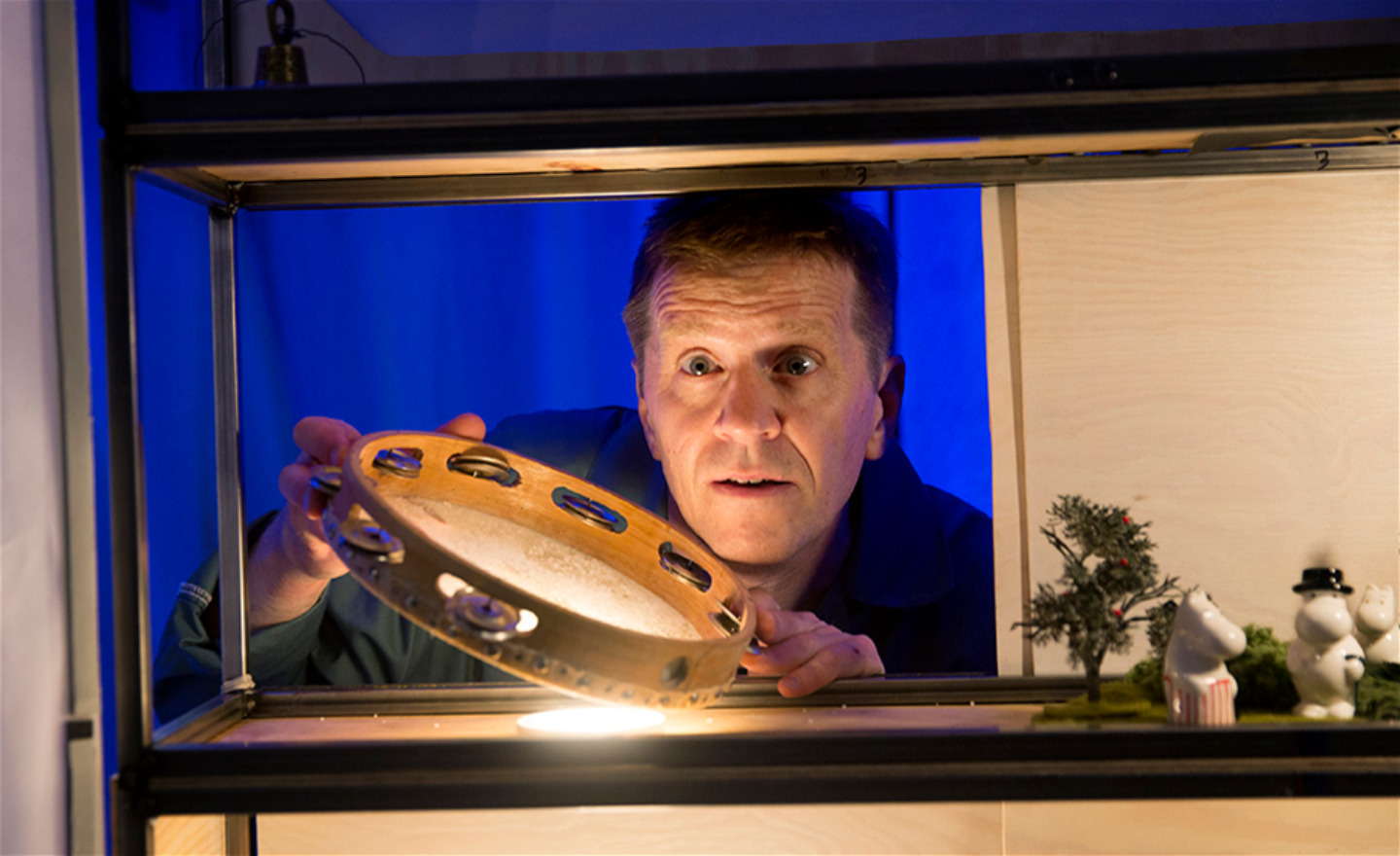 Skådespelare tittar ut genom lucka med tamburin, Bredvid porslinsfiguriner med muminfamiljen.