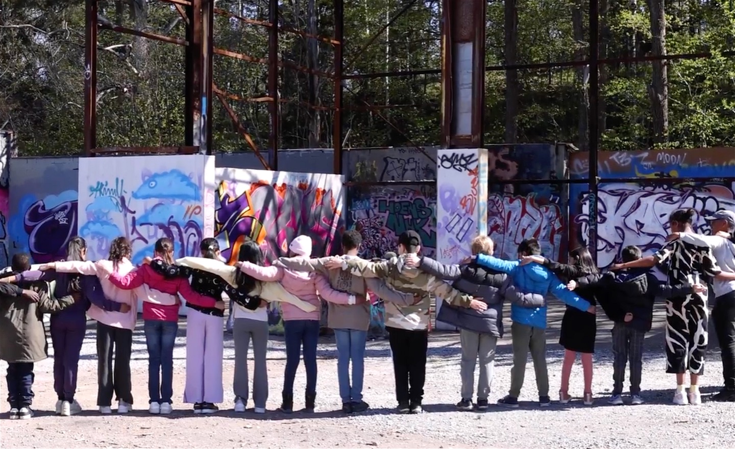 En skolklass med barn står med ryggen emot kameran och håller om varandras axlar. 