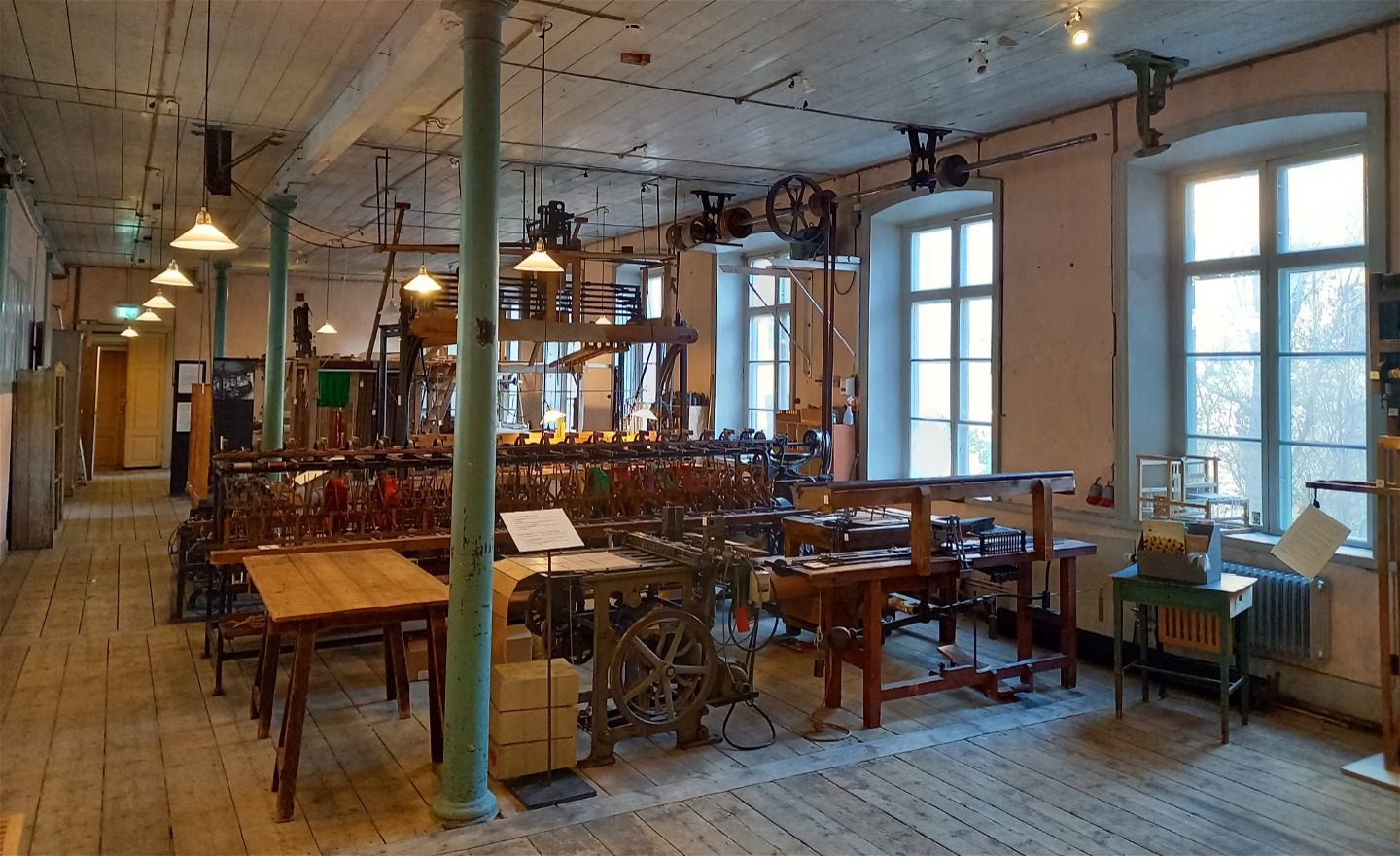 En stor fabrikssal med vävutrustning. rosa väggar, grönblå pelare och stora fönster.
