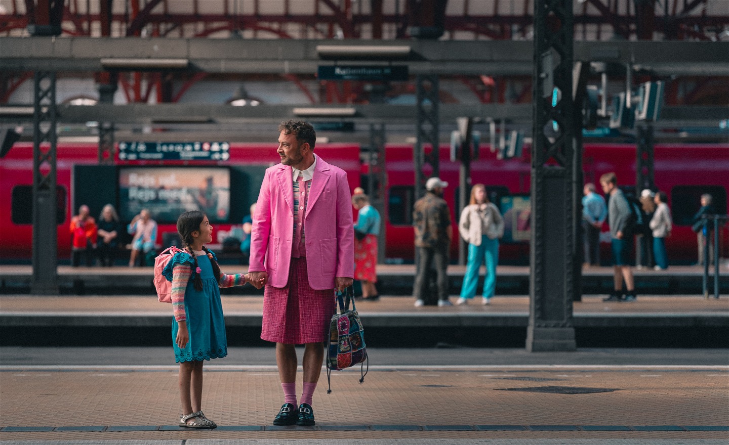 En flicka i blå klänning och rosa ryggsäck står vid en tågstation och håller en man i handen. Han är klädd i en rosa kavaj och kjol.
