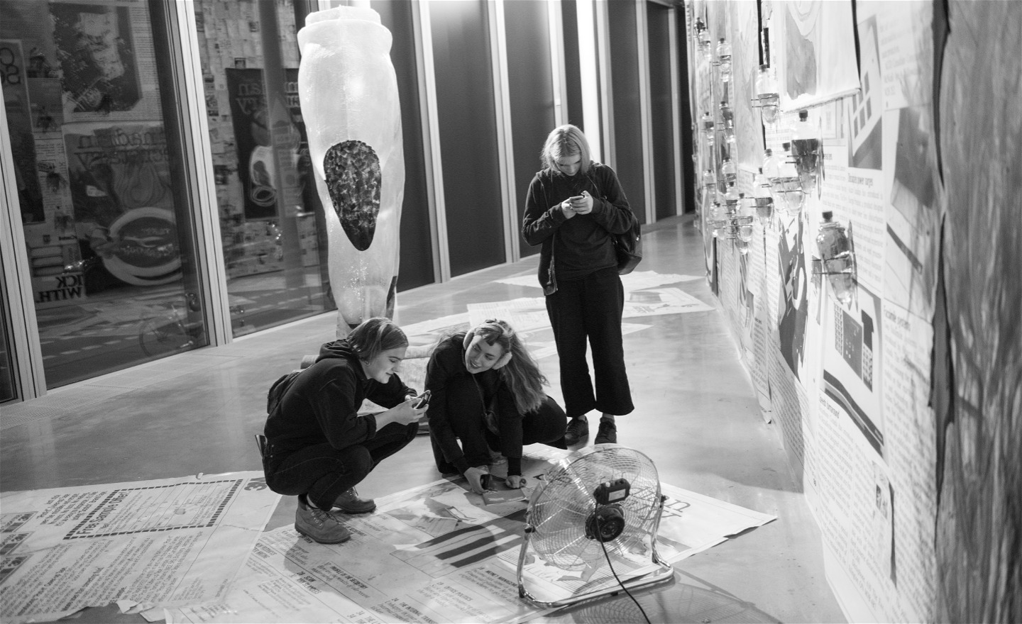 Svartvit bild av tre unga besökare som tittar på och fotar konstverk. 