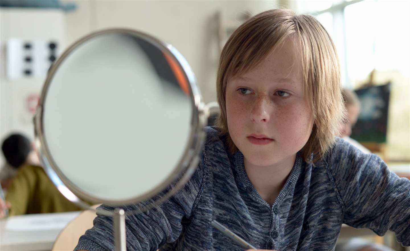 En pojke i mellanstadieåldern sitter i ett klassrum och betraktar sitt ansikte i en spegel.