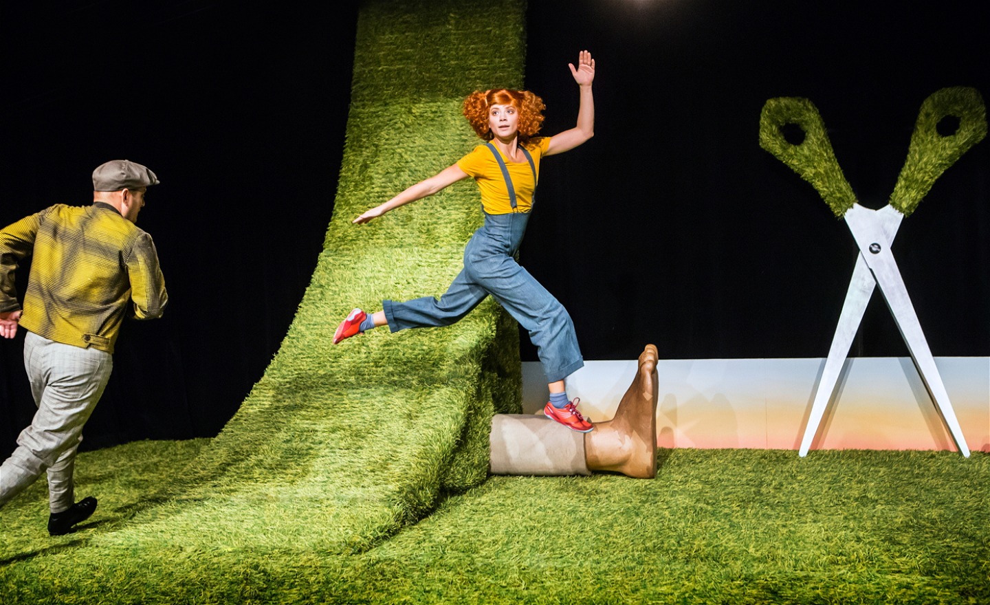 Två personer springer respektive hoppar på scenen som är klädd i konstgräs. På scenen finns också en sax större än en människa och en jättefot.