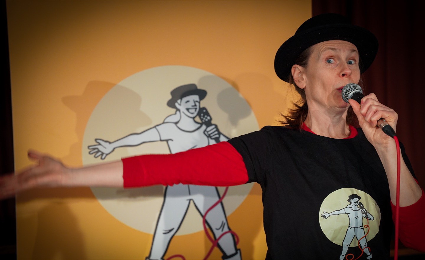 foto av Micaela som står upp med mikrofon, mot en saffransgul bakrund med en illustrerad bild av Micaela som står på samma sätt. På tröjan har även Micaela en miniatyr av samma bild. mikrofonsladden är röd