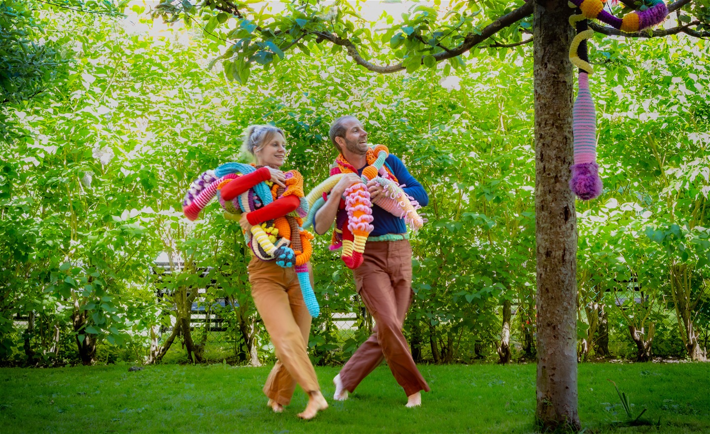 En kvinnlig och manlig dansare, dansar utomhus i  en park med famnen full av färglada fantasifulla virklingar