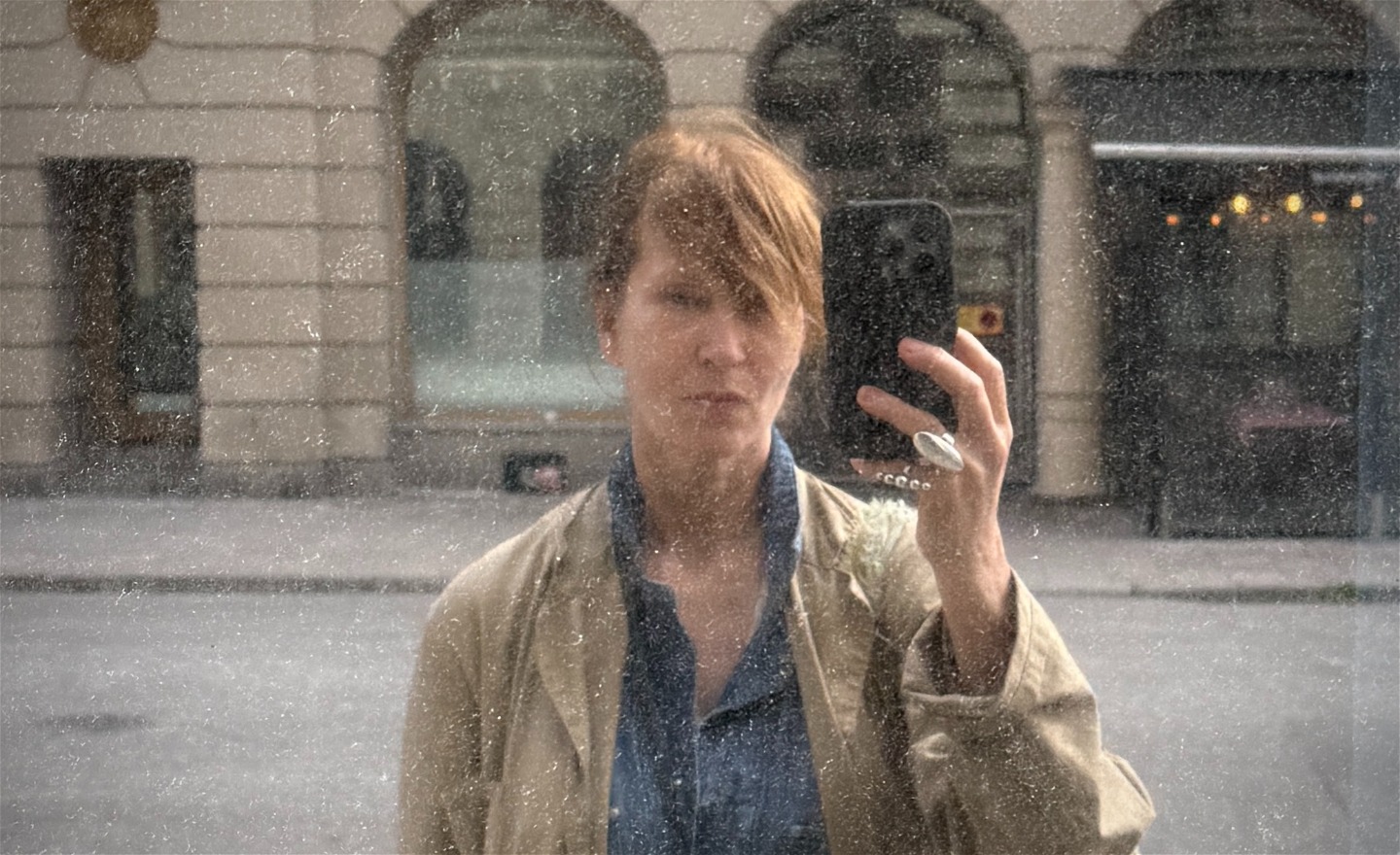 en närbild av en person med lång sidolugg som delvis täcker pannan. Bilden är tagen som en spegelselfie genom ett smutsig spegel, vilket ger en suddig effekt över ansiktet.