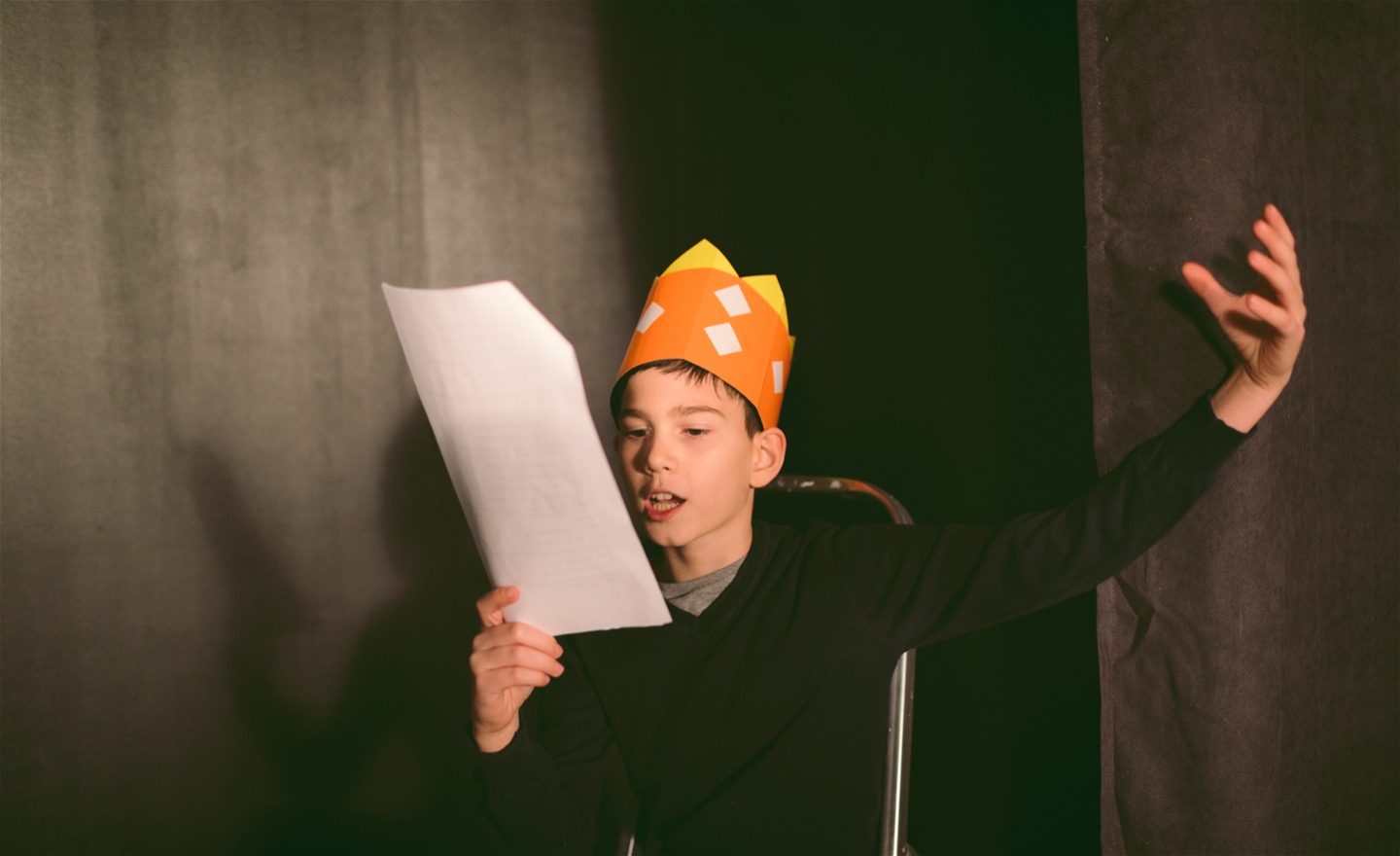 Pojke med en krona av papper på huvudet läser på ett papper och gör en gest med armen.