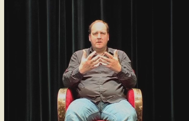 en man sitter på en stol framför ett svart draperi och pratar med händerna framför kroppen.