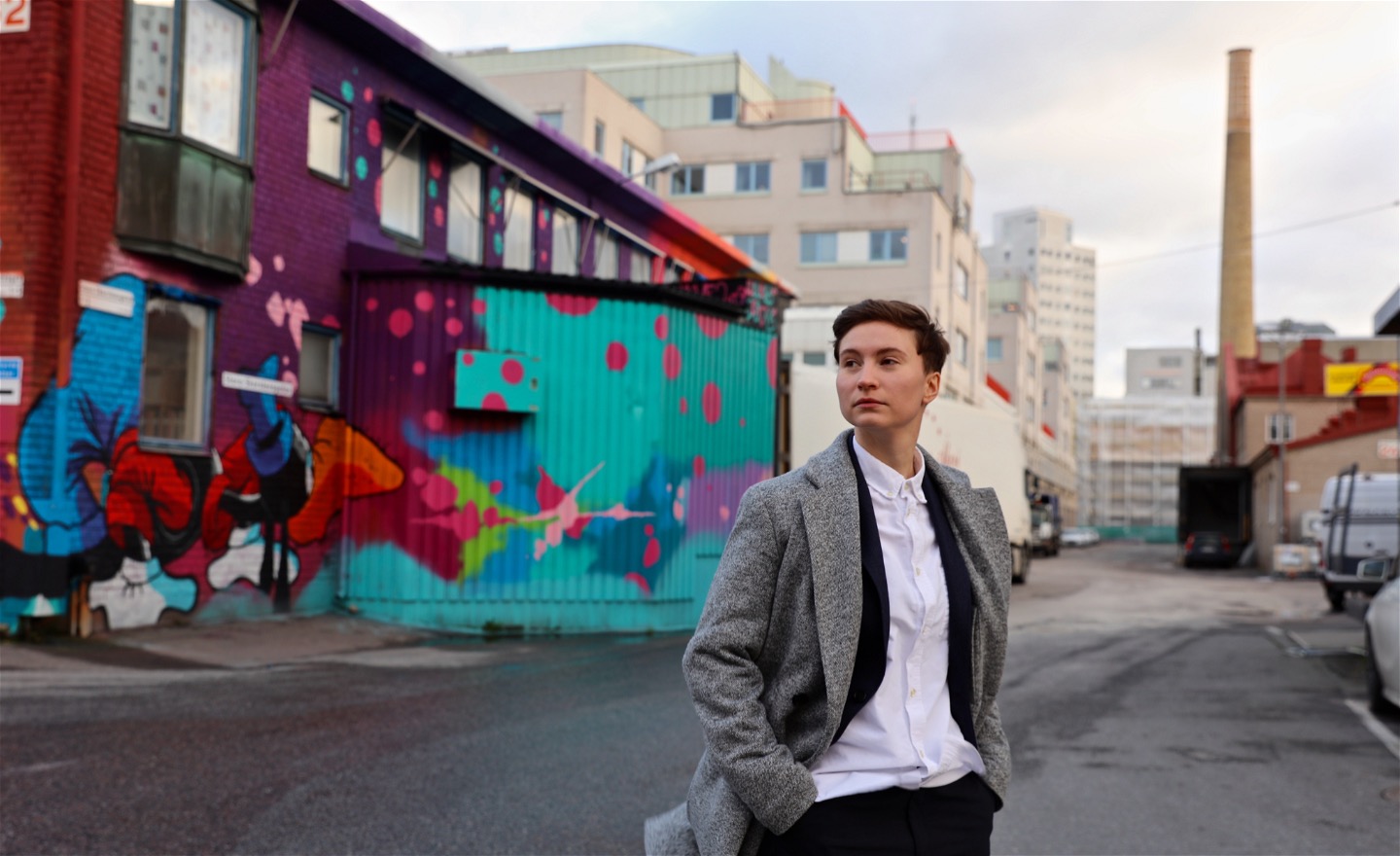 Sabina har på sig vit skjorta och rock, och står framför en grafittivägg