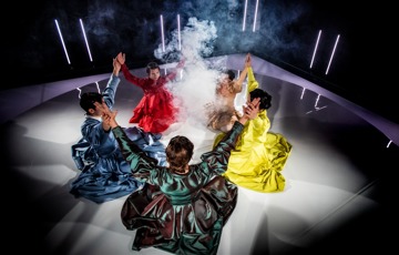 Fem dansare i färgstarka långklänningar i olika färger sitter i en ring och håller varandra i händerna med uppsträckta armar. I mitten stiger det upp rök.