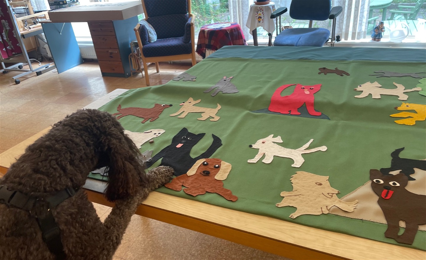 På ett bord ligger en stor bild gjord av textil. Den föreställer en äng med olika hundar som springer runt. De är urklippta i tyg och fastklistrade.  I vänstra hörnet av fotot finns en riktigt hund som böjer sig över bordet.