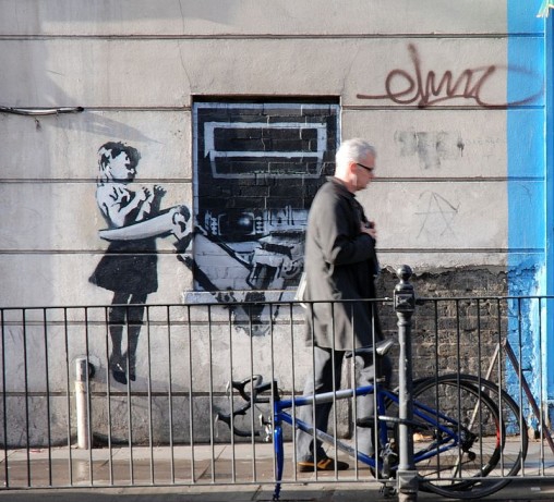 ett foto på en grafittimålning på en vägg. motivet är en en bankomat med klor som fångar in en liten flicka.