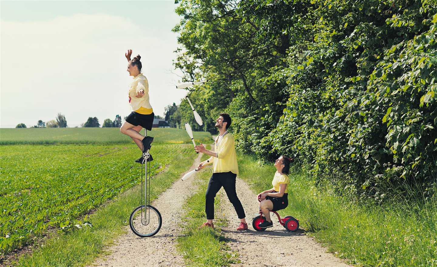Tre gulklädda cirkusartister utomhus. En på en högenhjuling, en jonglerandes med käglor och en på en röd liten trehjuling. 