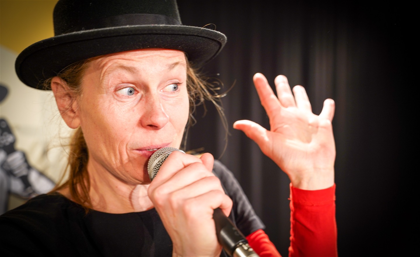 Micaela, med svart hatt pratar i med mikrofon, tittar underfundigt mot publiken och gör tittut-rörelse med ena handen.