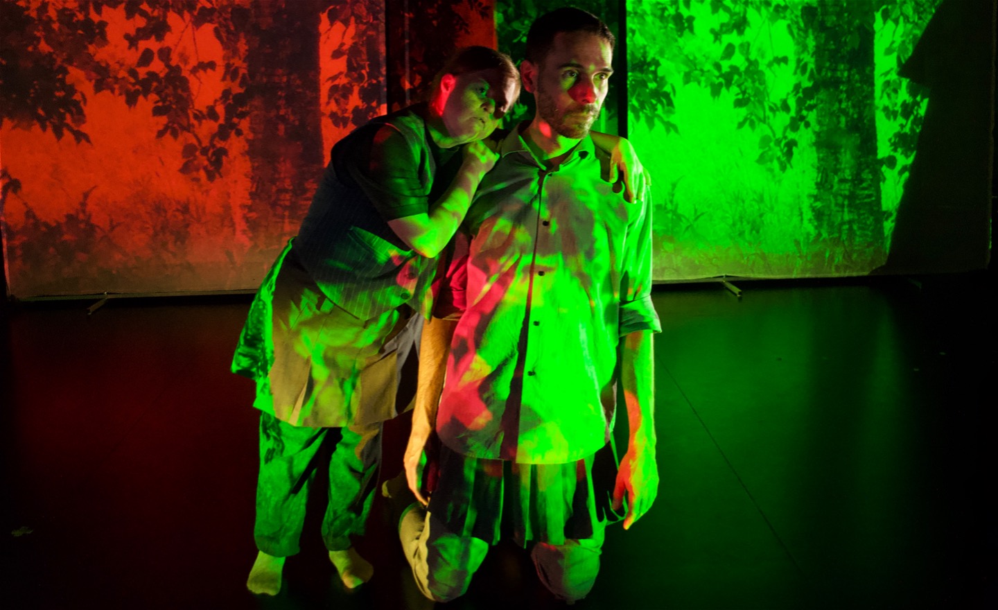 En dansare står på knä, en annan på fötterna och lutar sig emot, de är lika långa. Täckta av gröna och röda trädprojektioner. 