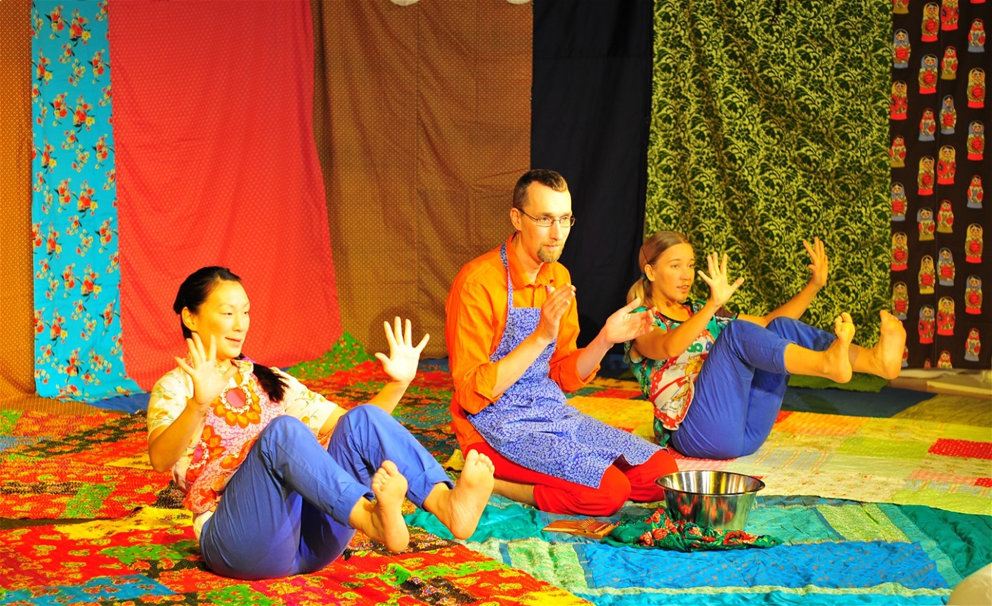 Tre dansare i färgglada kläder sitter på mönstrade mattor i starka färger och stäcker upp sina händer och fötter.