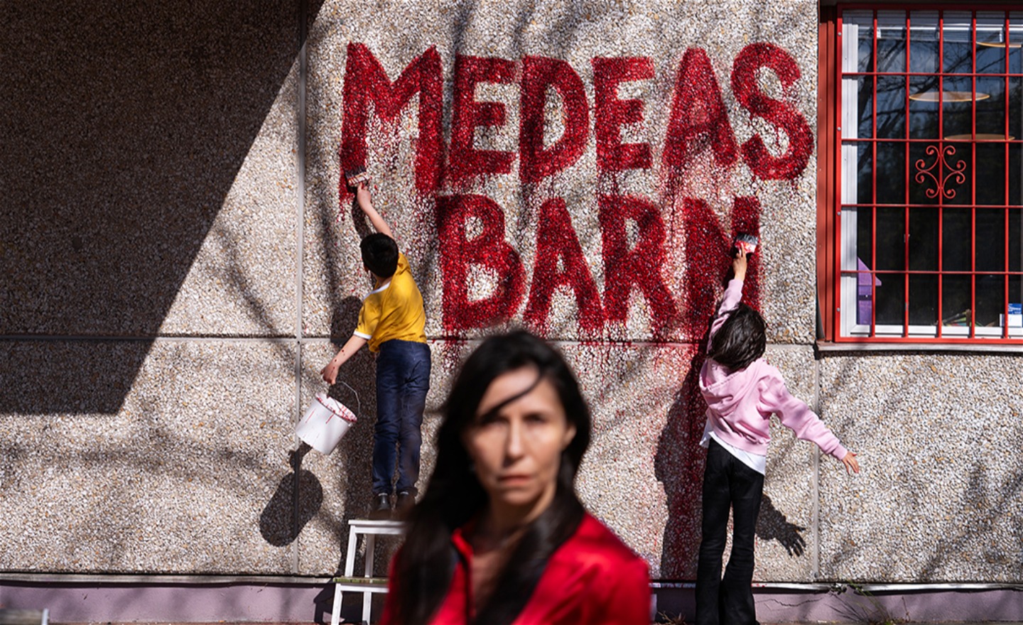 Tre tecknade figurer på en röd soffa utomhus vid SKHLM – en vuxen i mitten, ett hoppande barn med leksak och ett barn med en gul anka i knät.