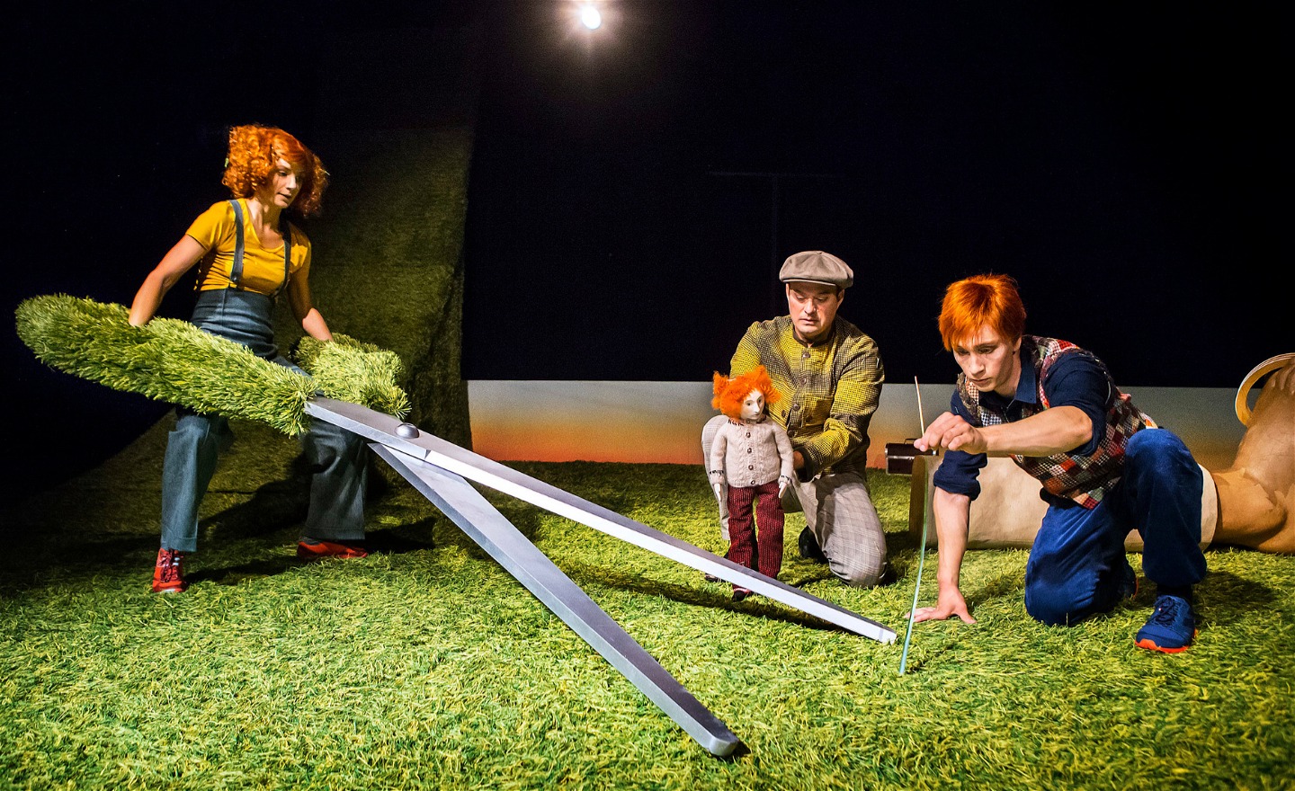 Tre personer är på en scen med konstgräs på golvet. Den ena håller i en sax större än en människa, den andra i en docka och den tredje i ett långt grässtrå.