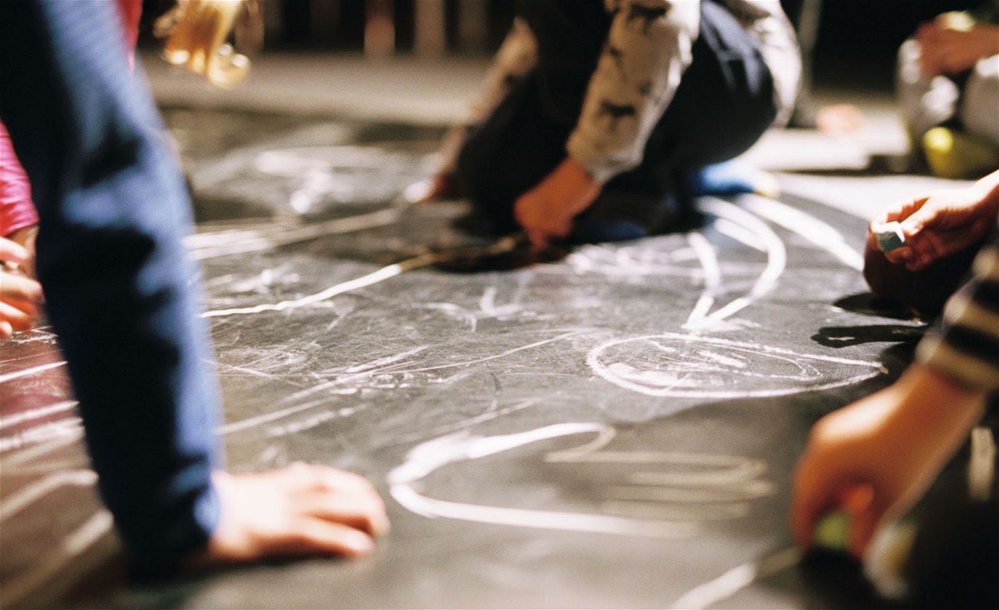 Barn sitter på ett gråsvart golv och ritar med vita kritor.