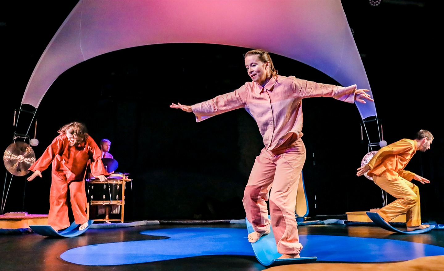 Tre dansare på balanserar på böjda brädor, en musiker spelar på slagverk